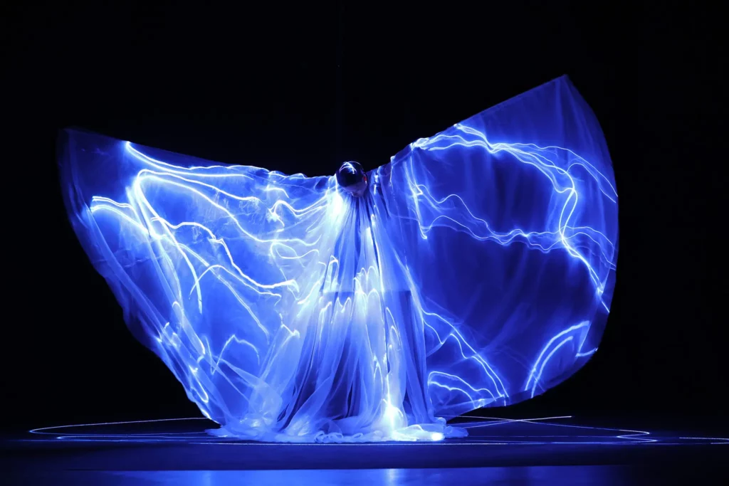 Performer on a dark stage wrapped in blue glowing fabric with neon light trails sweeping around.