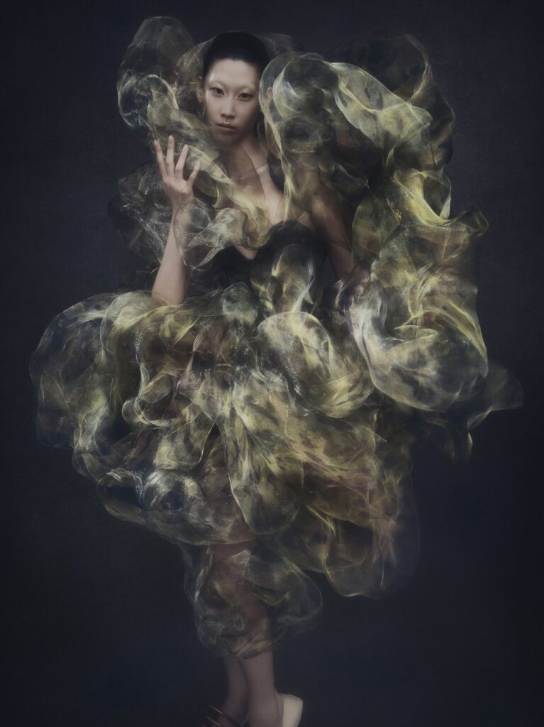 A person surrounded by billowing translucent yellow fabric against a dark background, posing dramatically.