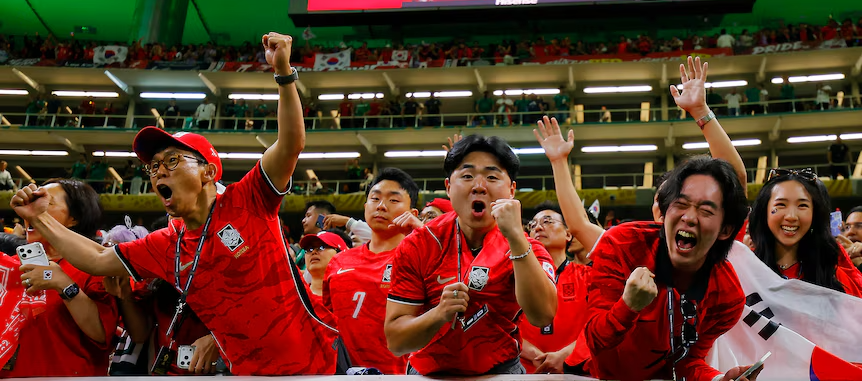 Group of excited fans in red jerseys cheering at a stadium, fists raised and mouths open in celebration amid a cheering crowd.