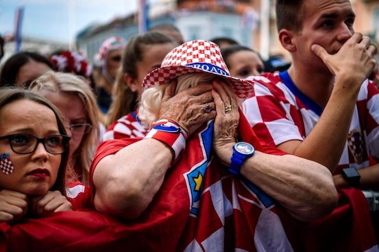 Older Croatian fan in a red-and-white checkered hat covers her face with hands amid a crowd of supporters, wearing red jerseys and flags