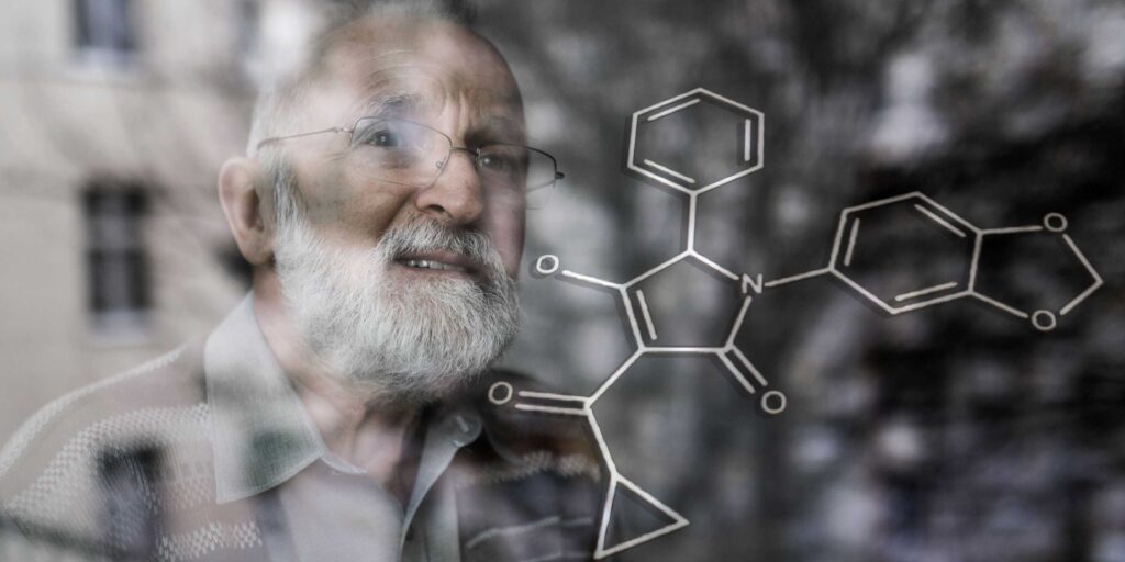 Older man with glasses and gray beard studying a chemical diagram on glass surface.