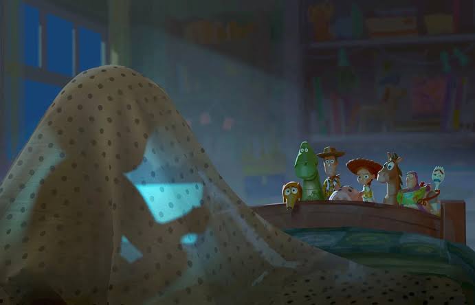 A child hides under a dotted blanket, peeking toward a row of toy figures on a curved shelf in a dim room, blue light on them.