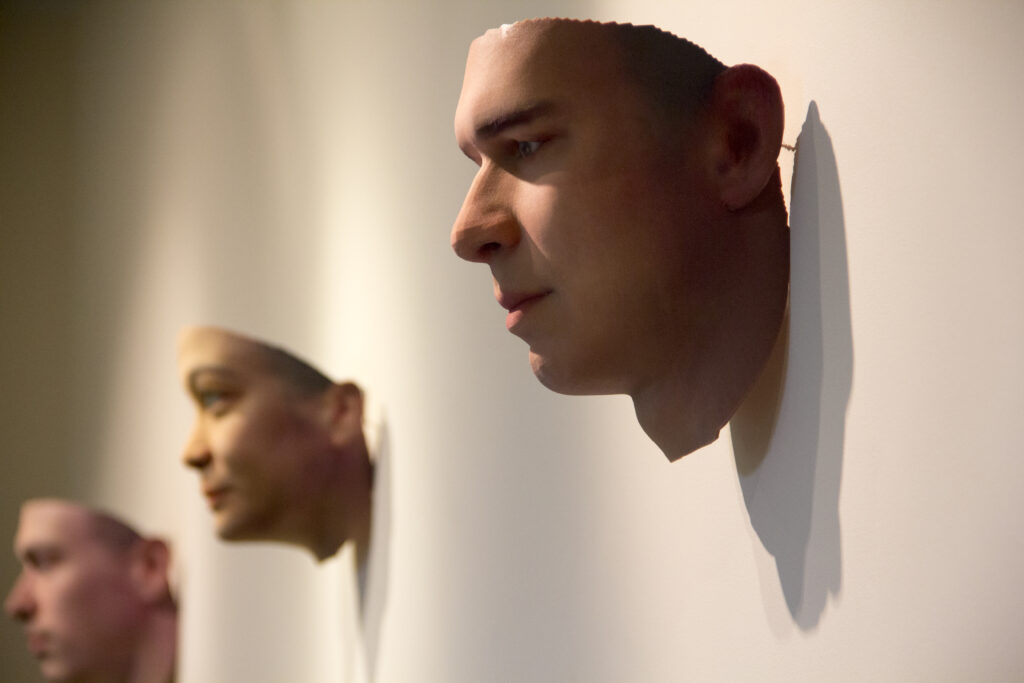 Heather Dewey-Hagborg's Stranger Visions - Lambda Biologics - Lambda Biologics Side profile of a life-size mannequin head mounted on a wall, with a soft shadow cast to the right in a gallery setting.