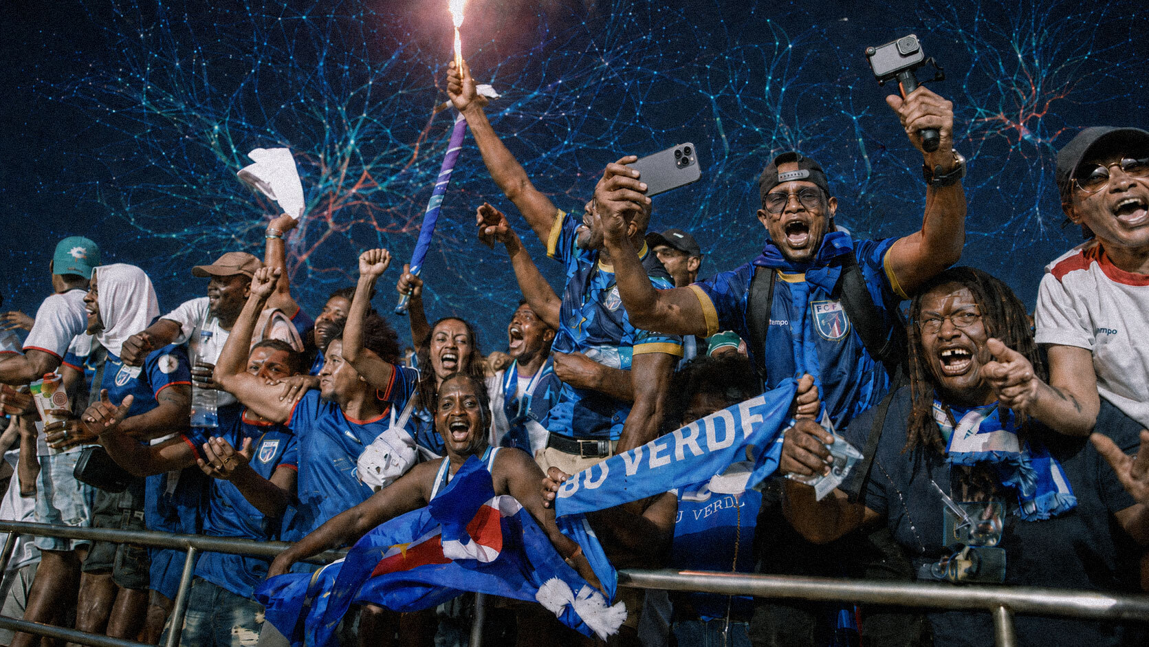 Group of ecstatic soccer fans in blue jerseys celebrating behind a railing, waving flags and filming with phones.