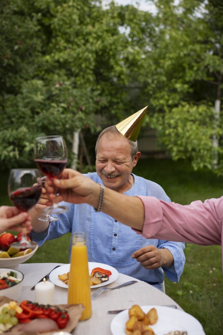 peoplecelebratingseniorbirthdaypartyoutdoorsgarden Lambda Biologics Lambda Biologics Man wearing a gold party hat toasts with a glass of red wine outdoors at a garden birthday dinner with friends nearby
