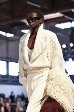 Runway model in a cream oversized knit cardigan with matching top and trousers, dark sunglasses, carrying a fluffy cream coat draped over her arm.